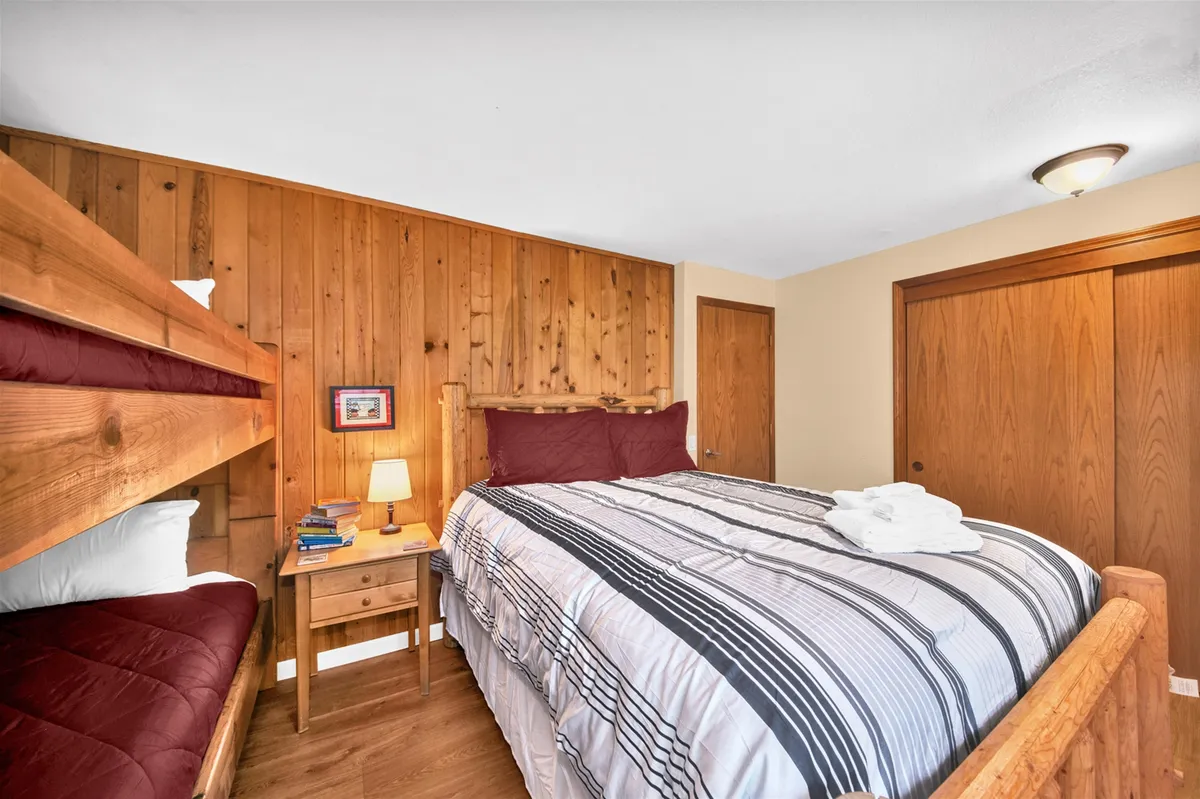 A welcoming downstairs bedroom featuring both a queen bed and a bunk bed, perfect for children, teens, or families. The wooden wall, nightstand with lamp, and closet doors create a playful yet cozy atmosphere ideal for group stays or story-filled nights.