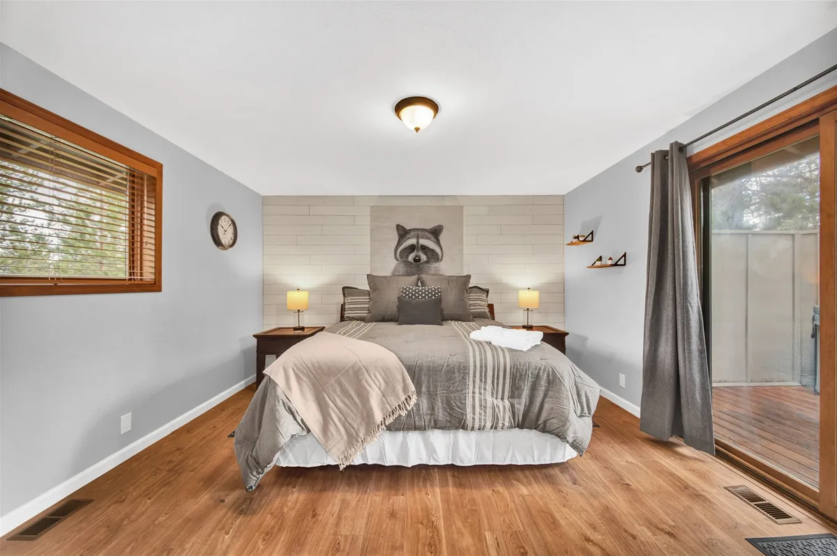A tranquil King bedroom with a comfortable bed dressed in decorative pillows, nightstands with lamps, and a wall art piece featuring a raccoon. The room is illuminated by natural light streaming through a sliding glass door that opens directly to the outdoors, suggesting convenient access to amenities like the hot tub.
