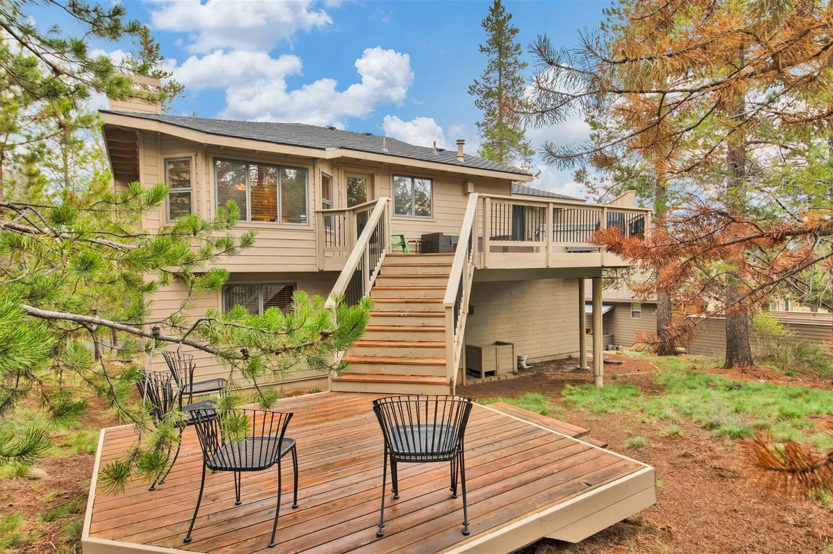 The deck features comfortable chairs and a staircase leading down into a wooded area, emphasizing the home’s connection to nature and providing space for outdoor gatherings. Sunlight filters through the trees, creating a warm and tranquil environment for families to gather and make memories.