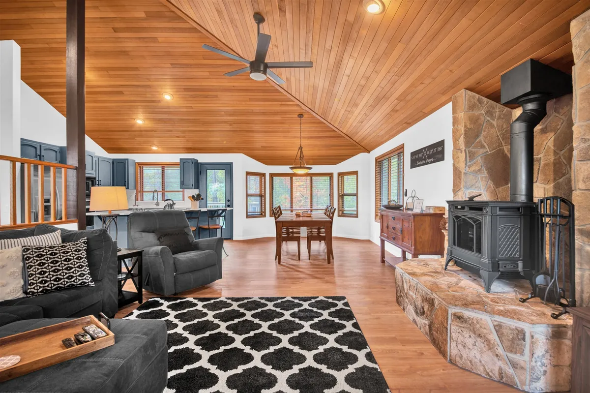 A welcoming open-plan living area with a striking stone fireplace, warm wooden ceilings, a well-set dining table, and plenty of comfortable seating. The layout is ideal for group gatherings, streaming movies, or simply unwinding by the fire with a beverage in hand.