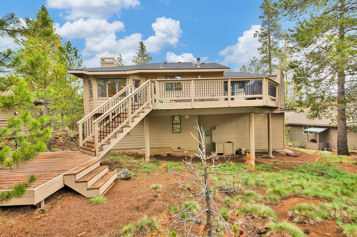 A spacious wooden deck set among trees, with stairs leading down to the yard and open sky above. The setting is tranquil and inviting, perfect for outdoor gatherings, relaxing after a day of activities, or family barbecues.