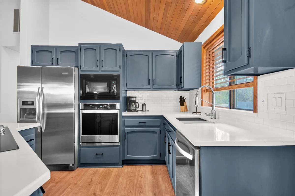 A close-up view of the modern kitchen, highlighting blue cabinetry, stainless steel appliances, white countertops, and a wooden ceiling. The spacious island with bar seating is visible, emphasizing functionality and style for cooking and entertaining.