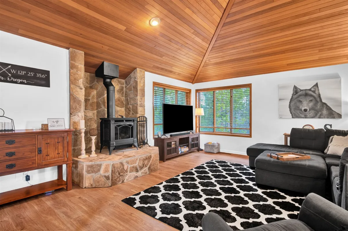 The image showcases the cozy living room of Tan Oak 2, featuring a striking stone fireplace as the centerpiece, flanked by large windows that let in natural light. Comfortable sofas invite guests to gather, while a black and white rug and wolf artwork add stylish, rustic charm. The overall atmosphere is warm, inviting, and ideal for relaxing or enjoying quality time with family and friends.