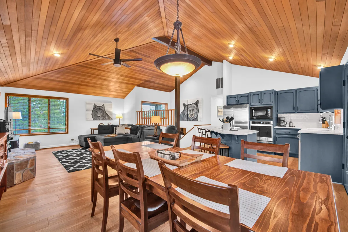 This image showcases the heart of the open-concept living space, featuring a spacious dining table, comfortable living area seating, a view into the kitchen area, and a warm wooden ceiling. The arrangement invites both relaxation and connection, with sightlines that tie the living, dining, and kitchen zones together.
