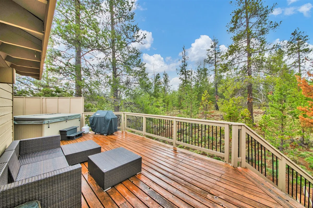 A spacious wooden deck bordered by a tree line, featuring an inviting hot tub and comfortable outdoor seating. The area is designed for relaxation and socializing, with ample space for guests to unwind after a day of adventure. The view through the trees hints at the surrounding natural beauty and resort environment.